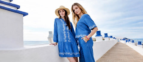 Two women in blue dresses with white embroidery standing on a white pathway by the sea.