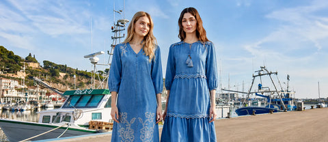 Two women in blue dresses standing by a marina with boats and buildings in the background.