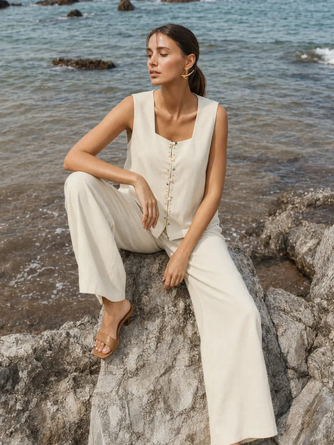 A woman sits on a rocky shoreline wearing a sleeveless cream-colored linen-like jumpsuit with wide-leg pants and tan flat sandals, with the ocean in the background.