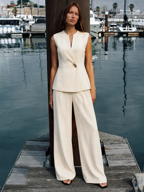 A woman stands on a wooden dock by the water wearing a sleeveless cream-colored two-piece suit with a structured peplum top and wide-leg trousers. The background shows a marina with boats and water.
