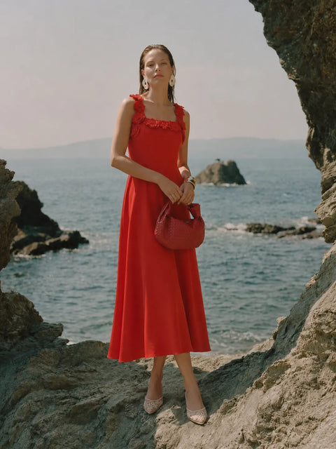 A woman stands on rocky coastal ground wearing a bright red midi dress with ruffled shoulder straps, holding a matching red handbag, with the ocean and sea stacks in the background.