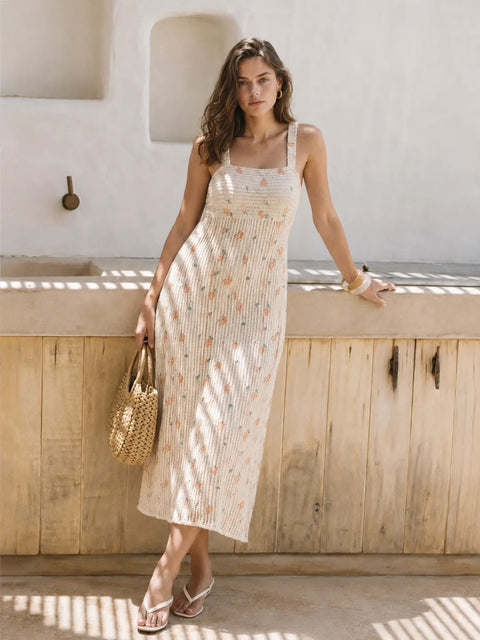 A woman models a sleeveless, square-neck midi dress with a cream base and a delicate, small floral print in soft peach and muted green. She is standing outdoors in front of a rustic wooden counter and white stucco wall, holding a woven straw bag.