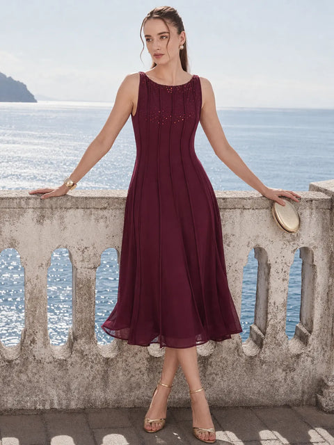 A woman stands outdoors by a stone railing overlooking the ocean, wearing a sleeveless midi dress in deep burgundy with subtle beaded detailing at the neckline. She is holding a small round clutch and wearing metallic heeled sandals.