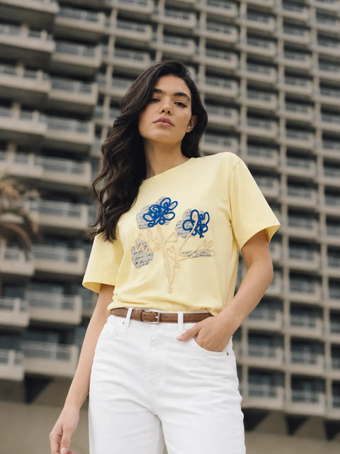A woman wearing a pale yellow short-sleeve t-shirt with blue floral embroidery, paired with white pants and a brown belt, standing in front of a large concrete apartment building.
