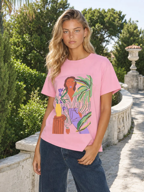 A woman wearing a pink graphic t-shirt with a colorful illustration of a woman and plants, paired with dark blue jeans, standing outdoors on a stone walkway surrounded by green trees.