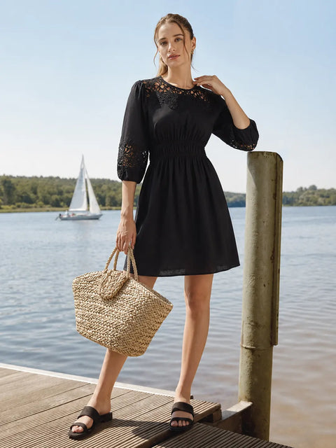 A woman stands on a wooden dock by the water wearing a black dress with lace detailing on the bodice and sleeves, holding a woven straw tote bag. A sailboat is visible in the background on the water under a clear blue sky.