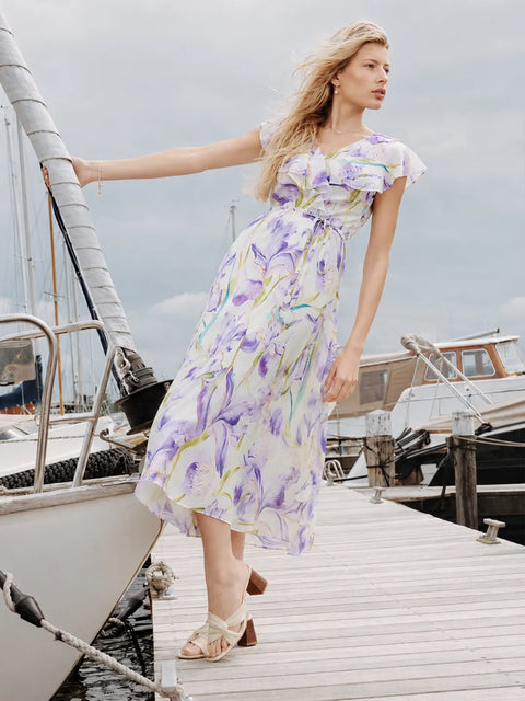 A woman wearing a white midi dress with a purple and green floral print stands on a wooden dock next to a boat, holding onto the mast.