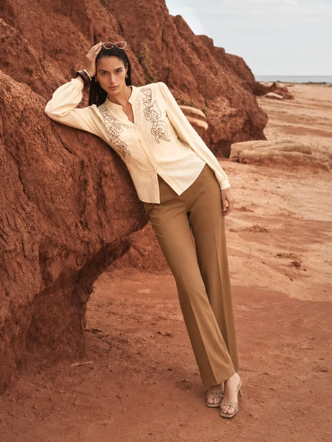 A woman posing outdoors against a large reddish-brown rock formation on a sandy beach, wearing a cream embroidered blazer, tan trousers, and metallic sandals.